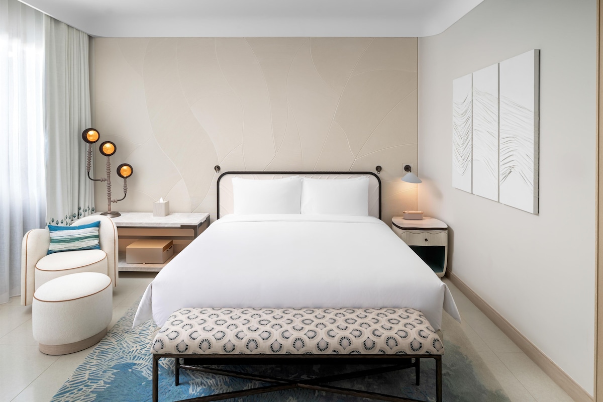 A hotel room with a bed, bedside tables, a desk, a chair, and a bench.