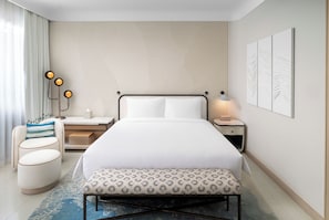 A modern hotel room with a large bed, bedside tables, a bench, and a patterned rug.