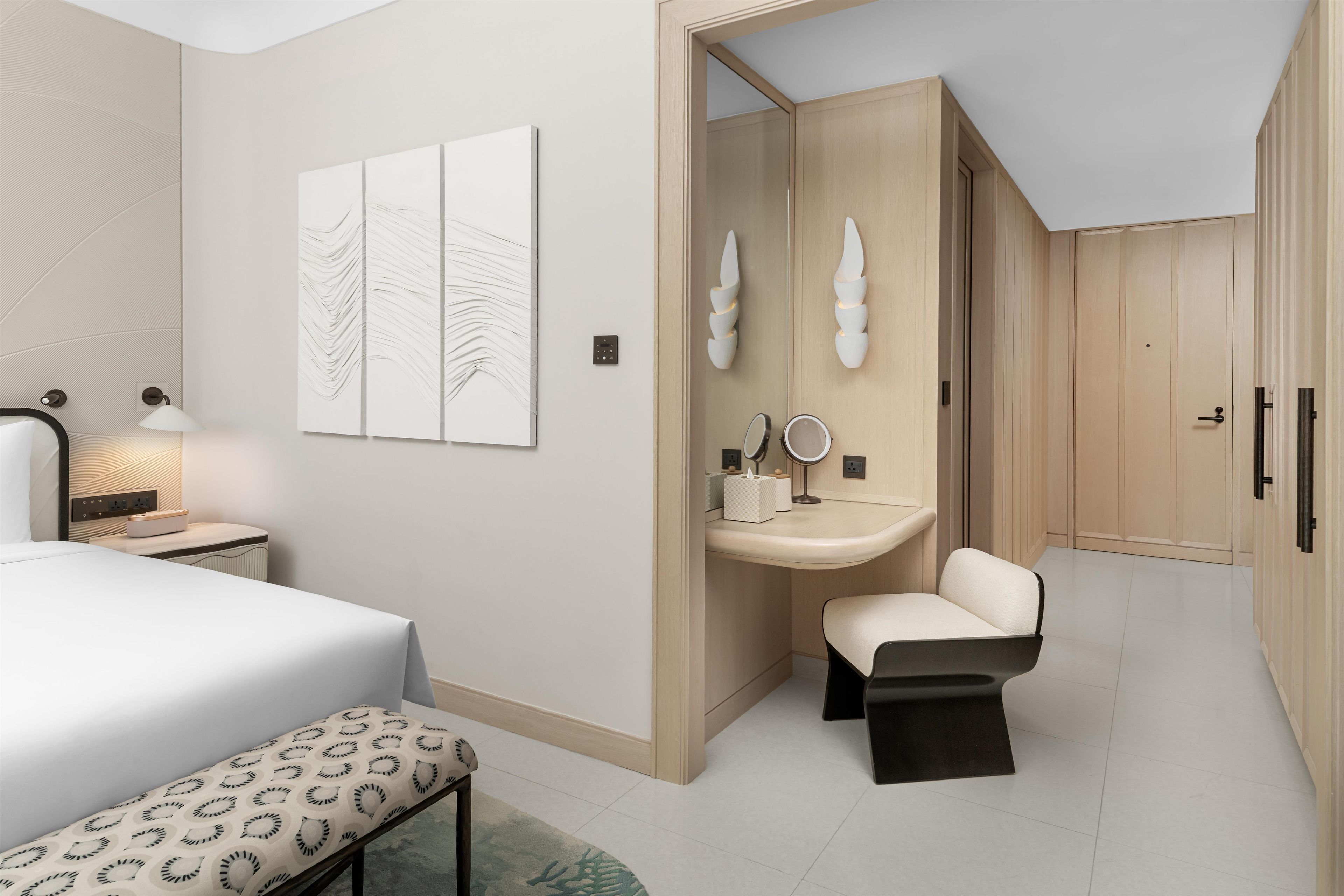 A modern bedroom with a bed, bedside table, lamp, artwork, vanity with mirror, and a bench.