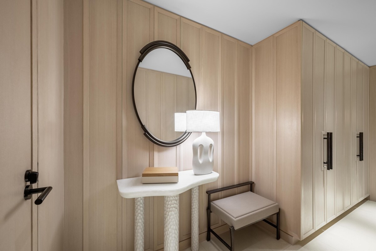A room with wooden panel walls, a white console table with a book and a lamp, a small bench, a mirror, and a door with a handle.