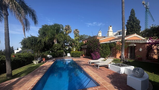 A rectangular swimming pool with a tiled deck, surrounded by palm trees and a building with a tiled roof. - Luxury Villa with All Amenities in Marbella   – Pool, Jacuzzi, Barbecue, Wood-Fired Oven (Benahavís)
