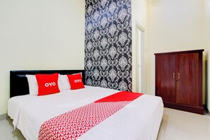 Una cama con colcha blanca y roja, dos almohadas rojas con el texto 'OYO', un papel tapiz estampado y un armario de madera.