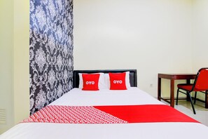 Habitación de hotel con una cama, dos almohadas rojas con el texto 'OYO', una cortina estampada, un escritorio de madera y una silla roja.