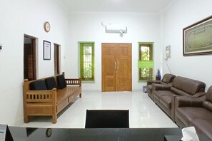 Uma sala de estar com um sofá de madeira, um sofá de couro marrom, um banco de madeira, uma porta com um vitral verde e um ar condicionado fixado na parede.