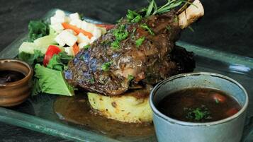 A plate with a roasted lamb leg, mashed potatoes, a side salad, and two small bowls of sauce.