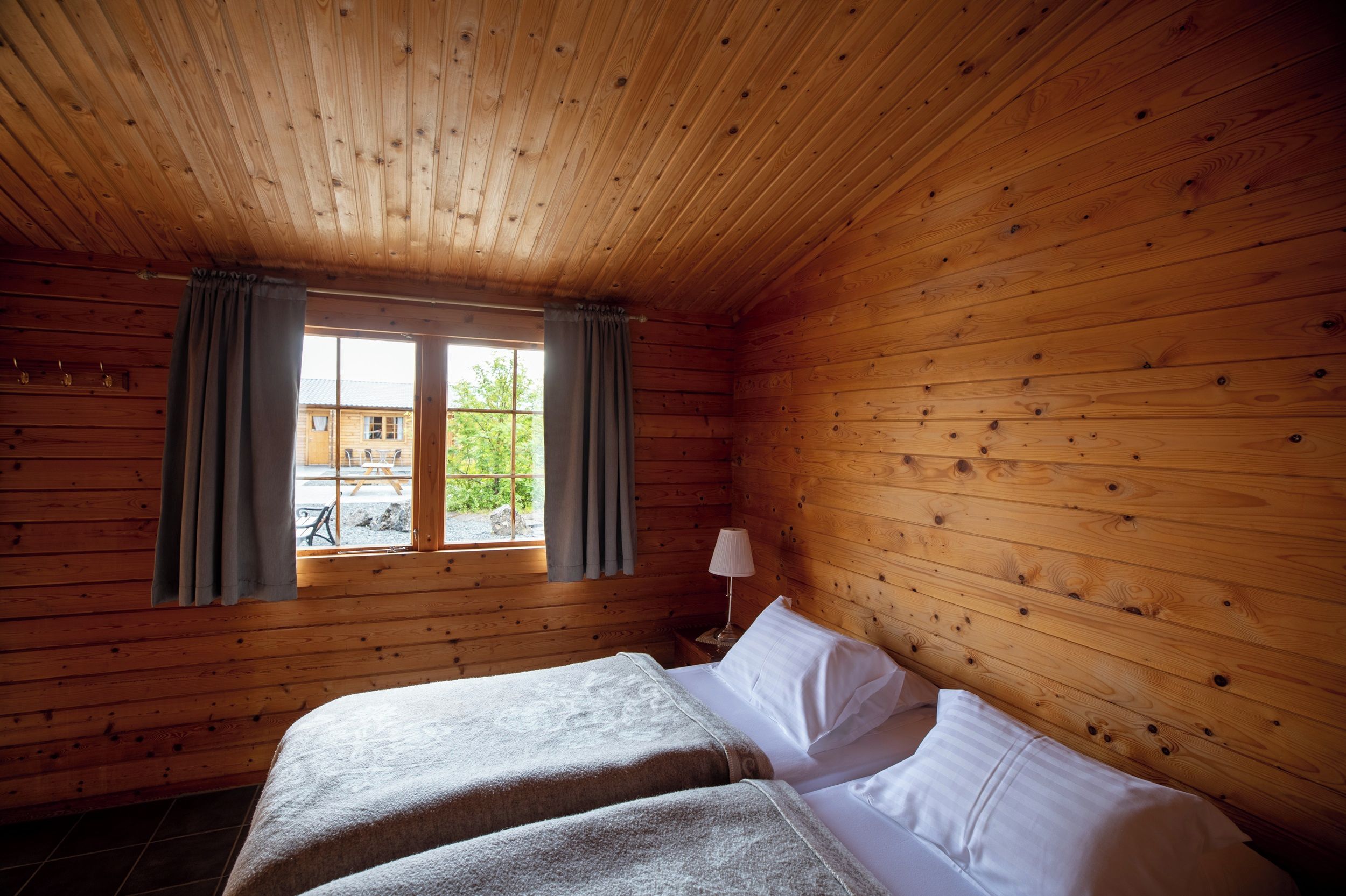 A wooden cabin room with a bed, a window with curtains, and a lamp on a bedside table.