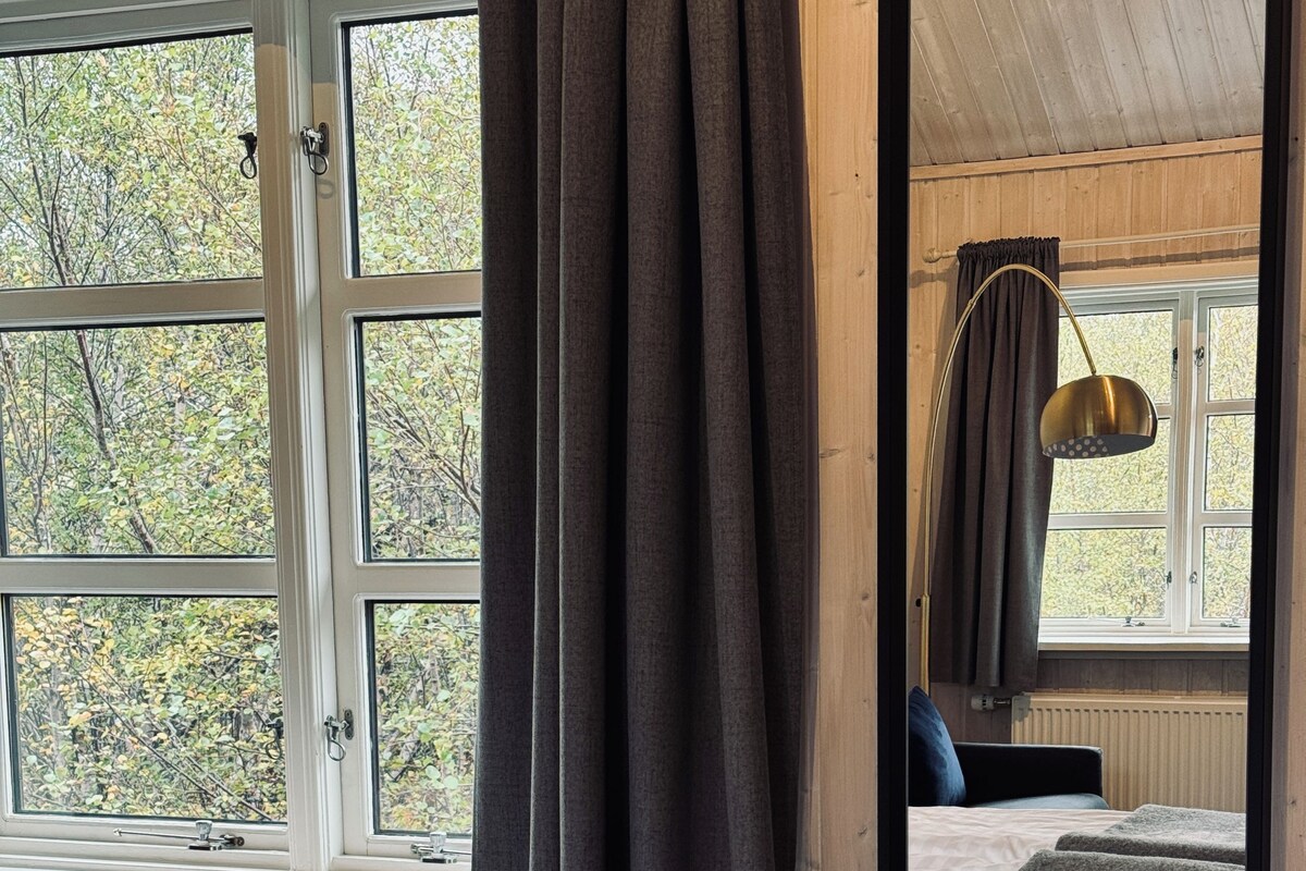 Un dormitorio con un ventanal grande, una cama con colcha estampada, una mesita de noche y un espejo que refleja la habitación.