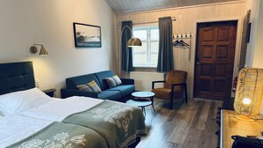 A hotel room with a bed, a sofa, a chair, a small table, a lamp, a window with curtains, a coat rack, and a door.