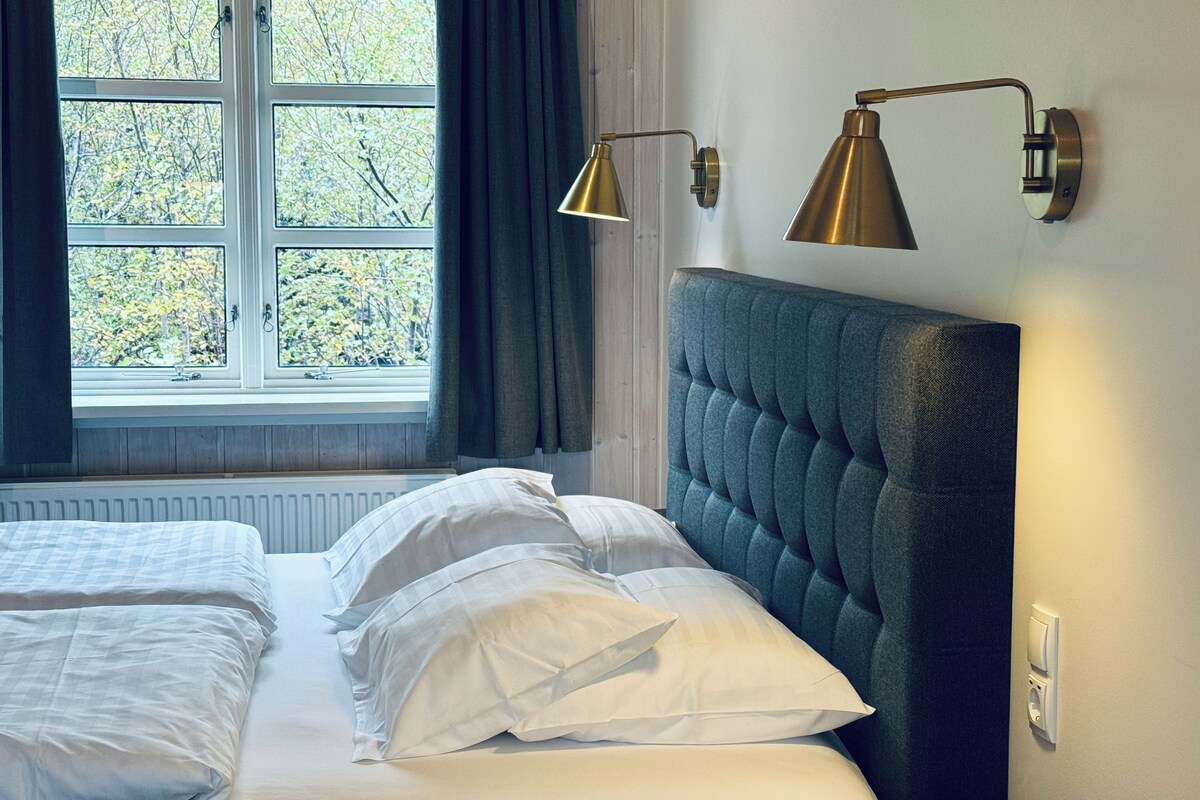 A bedroom with a bed, two bedside lamps, a window with curtains, and a radiator.