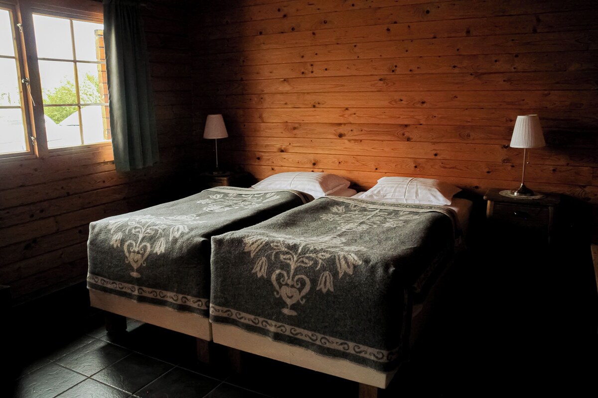 Ein Zimmer mit Holzwänden, zwei Betten mit passenden Kopfteilen, einem Fenster mit Vorhängen und einem Nachttisch mit Lampe.