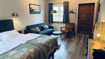 A hotel room with a bed, a sofa, a chair, a small table, a lamp, a window with curtains, a coat rack, and a door.