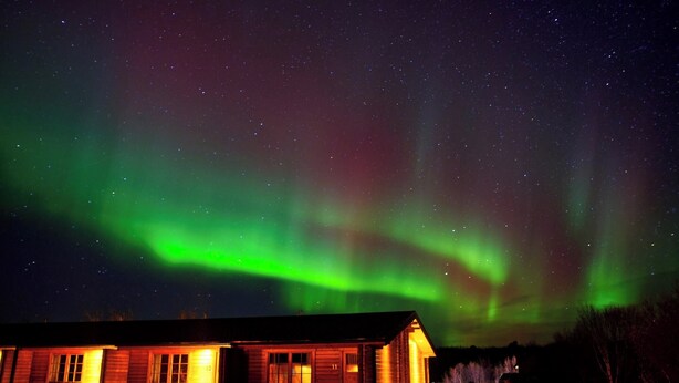 A wooden cabin with a porch, under a starry night sky with visible aurora borealis.