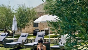 Un área de piscina con sillas de descanso y sombrillas, rodeada de follaje y edificios de piedra.