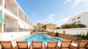 A swimming pool with lounge chairs, a white building with balconies, and a clear sky. - Hostal S Algaret (Mercadal)