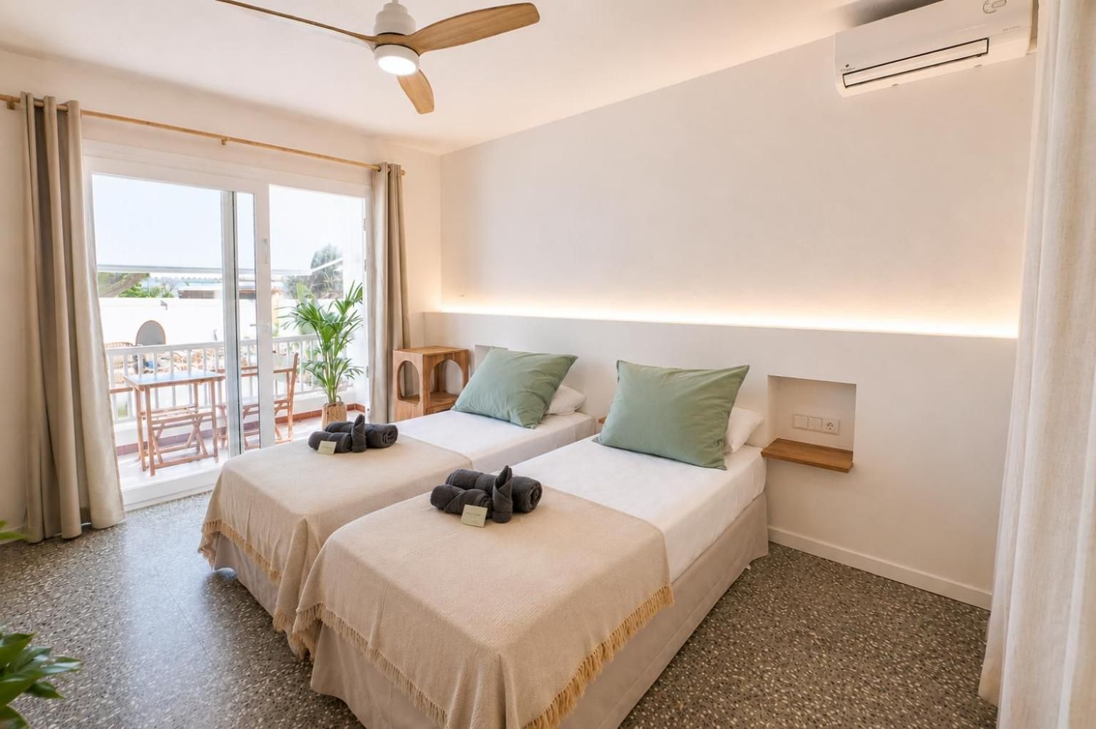 Quarto de hotel com duas camas, um ventilador de teto, uma mesinha e vista para o exterior.