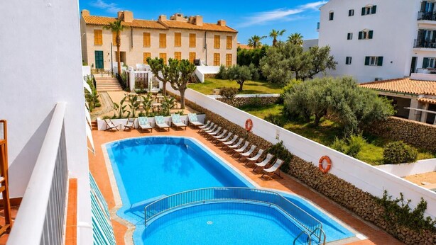 A rectangular swimming pool with a tiled deck, surrounded by lounge chairs and a white wall with a stone border. - Hostal S Algaret (Mercadal)
