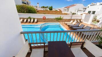 Área de piscina com espreguiçadeiras e uma mesa, cercada por edifícios brancos e um céu limpo.