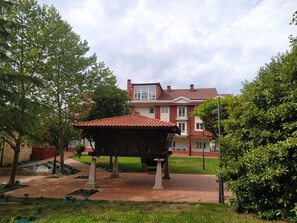 Ein Gazebo mit Ziegeldach, umgeben von einem gepflasterten Bereich mit Steinpfeilern, vor einem rotziegelbau mit Balkon.