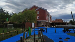 A red brick building with a flat roof, surrounded by a blue play area with wooden structures and a rope bridge. - Hotel La Campana (Llanera)
