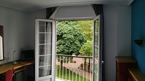 Et værelse med en seng, et skrivebord med en computer, en stol og et åbent vindue med udsigt til grønne omgivelser.