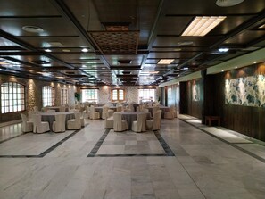 Un amplio salón de banquetes con varias mesas redondas y sillas, paredes de piedra y grandes ventanales.