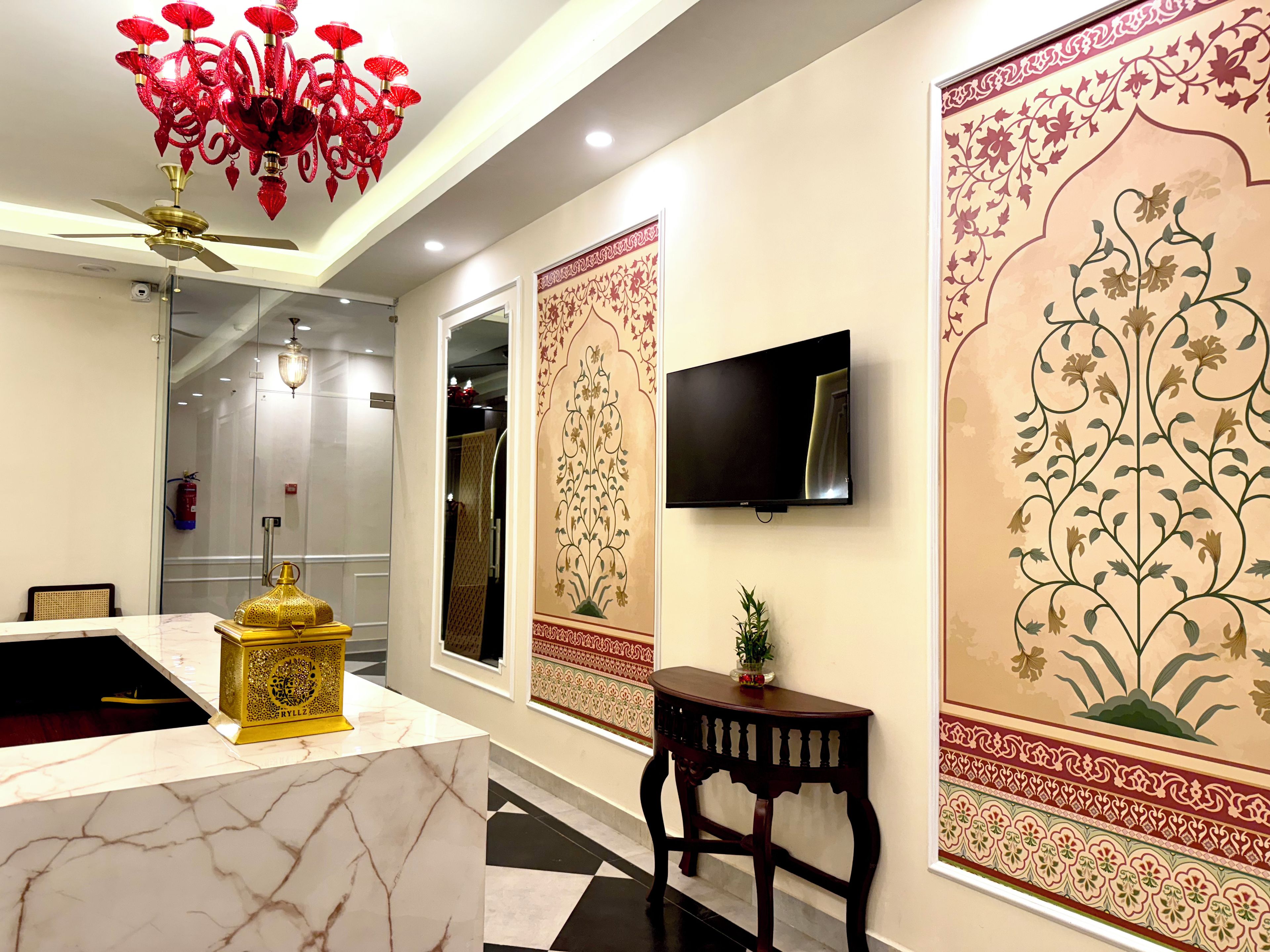 A hotel lobby with a marble reception desk, a golden lamp, a red chandelier, a flat-screen TV, and ornate wall decorations.