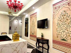 Un vestíbul d'hotel amb una taulell de recepció de marbre, una làmpada daurada, un penjador de cristall vermell, un televisor de pantalla plana i decoracions murals ornamentades.