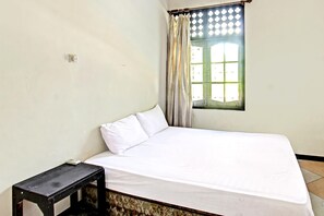 Una cama individual con ropa de cama blanca, una mesita de noche pequeña, una ventana con cortina y un enchufe de pared.