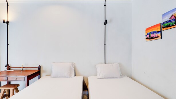 Una habitación pequeña y limpia con dos camas individuales, un escritorio de madera y una silla. Hay dos lámparas fijadas a la pared y un cuadro pequeño.