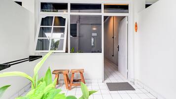 A clean, modern interior with white walls, a tiled floor, and a large window with curtains. There are two wooden stools and a potted plant in the foreground.