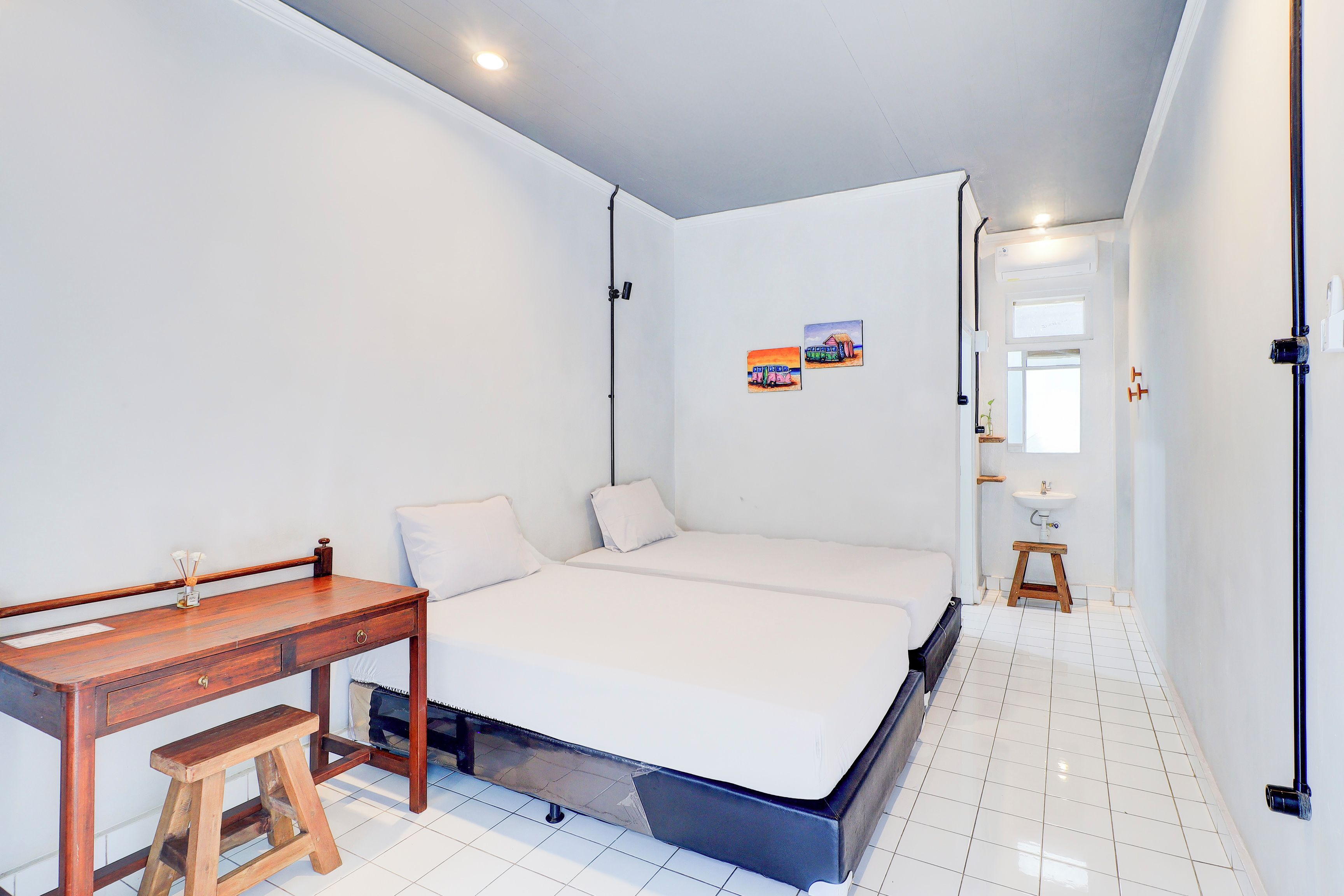 Een slaapkamer met een bed, een bureau en een klein krukje. Er is een raam met uitzicht op een gebouw en een schilderij aan de muur.