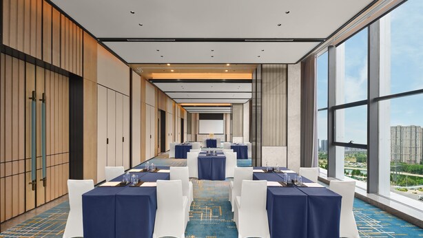 Un salón de conferencias con mesas dispuestas para una reunión, amplias ventanas que ofrecen vistas a la ciudad y un diseño interior moderno.
