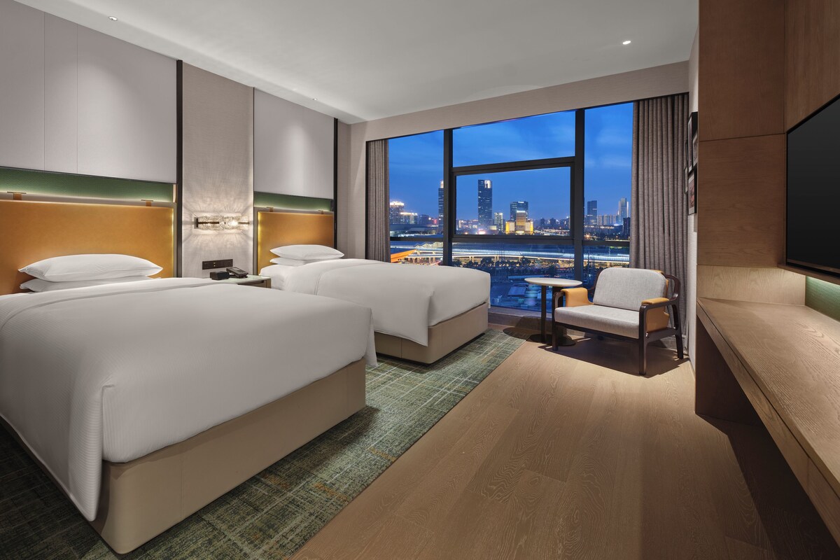 A hotel room with two beds, a seating area, a TV, and a city view.