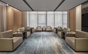 Een hotelkamer met een groot raam, een blauw tapijt en beige banken rond een centrale tafel.