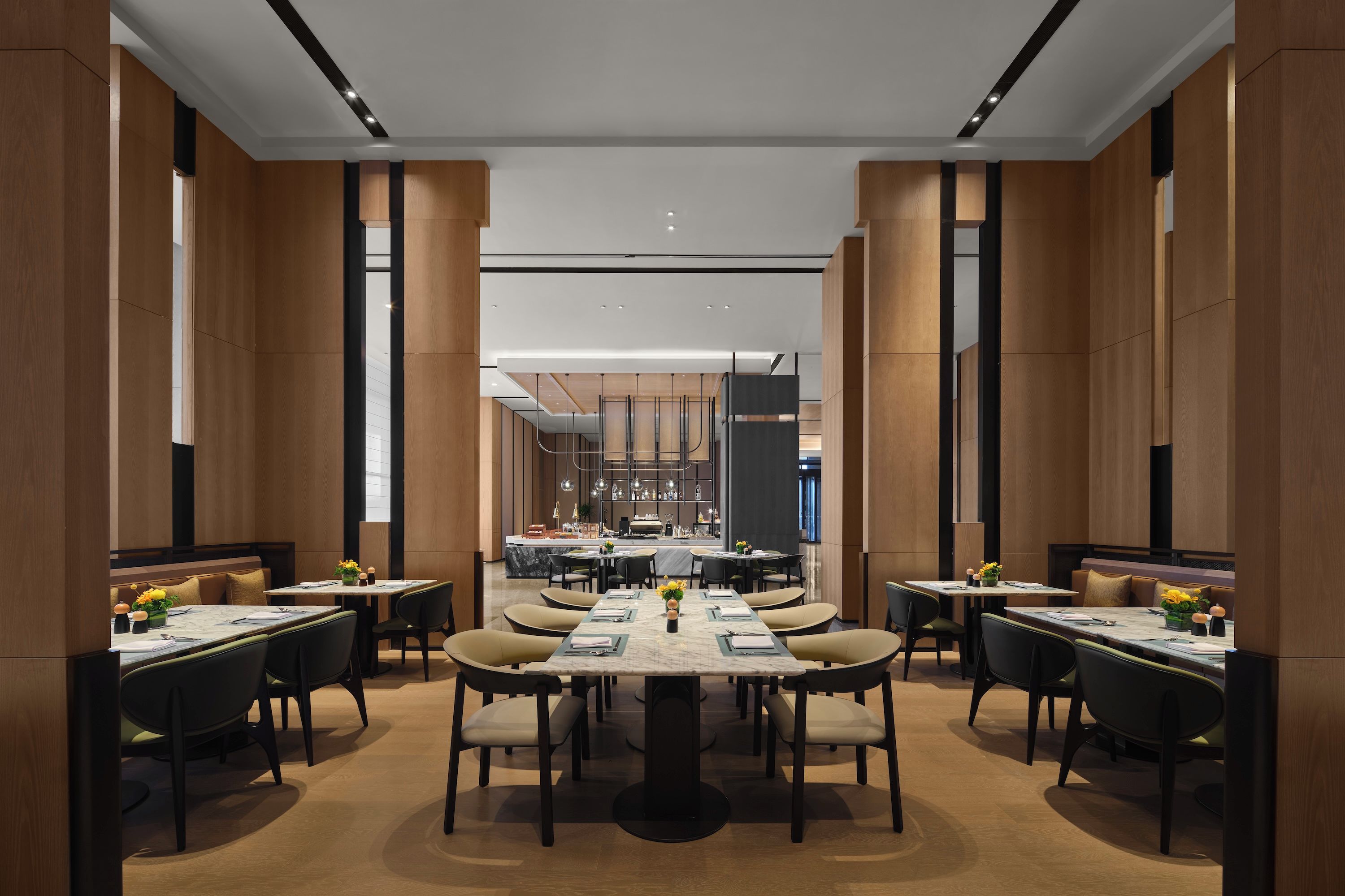 Un comedor moderno con techos altos, paneles de madera y una barra central. Las mesas están dispuestas con sillas, y el espacio está bien iluminado.