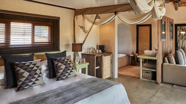 A bedroom with a bed, pillows, a nightstand, a lamp, a window with blinds, a bathroom with a sink and mirror, and a staircase leading to another room. - Toro River Lodge (Kareeberg)
