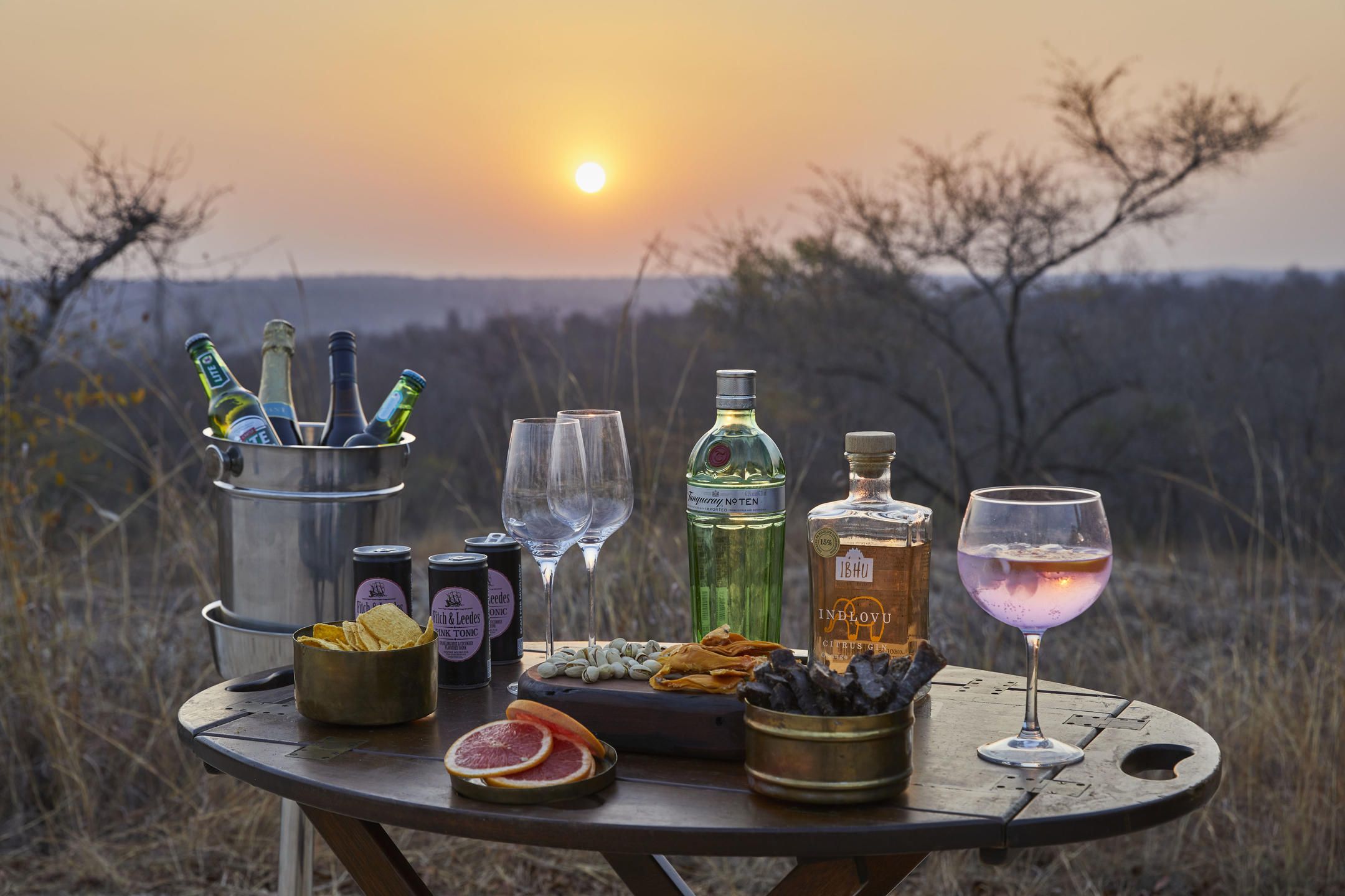 A table set outdoors with bottles, glasses, snacks, and a sunset view.