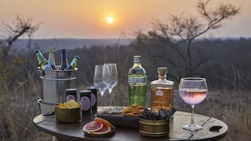 A table set outdoors with bottles, glasses, snacks, and a sunset view.
