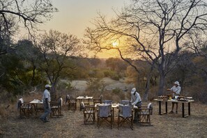 Un área de comedor al aire libre con mesas y sillas, rodeada de árboles y una puesta de sol al fondo.