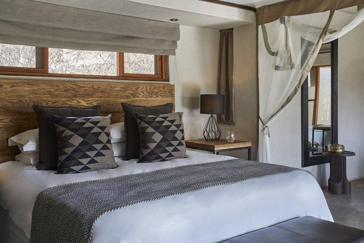 A bedroom with a large bed, a bedside table with a lamp, a window with curtains, and a view of trees outside.
