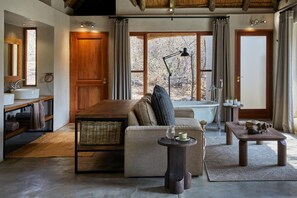 A cozy living room with a sofa, a small table, a lamp, and a view of the outdoors through large windows.