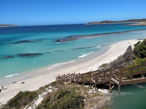 Ein Strand mit klarem türkisblauem Wasser, einem sandigen Ufer und einer Holztreppe, die zum Strand hinabführt.