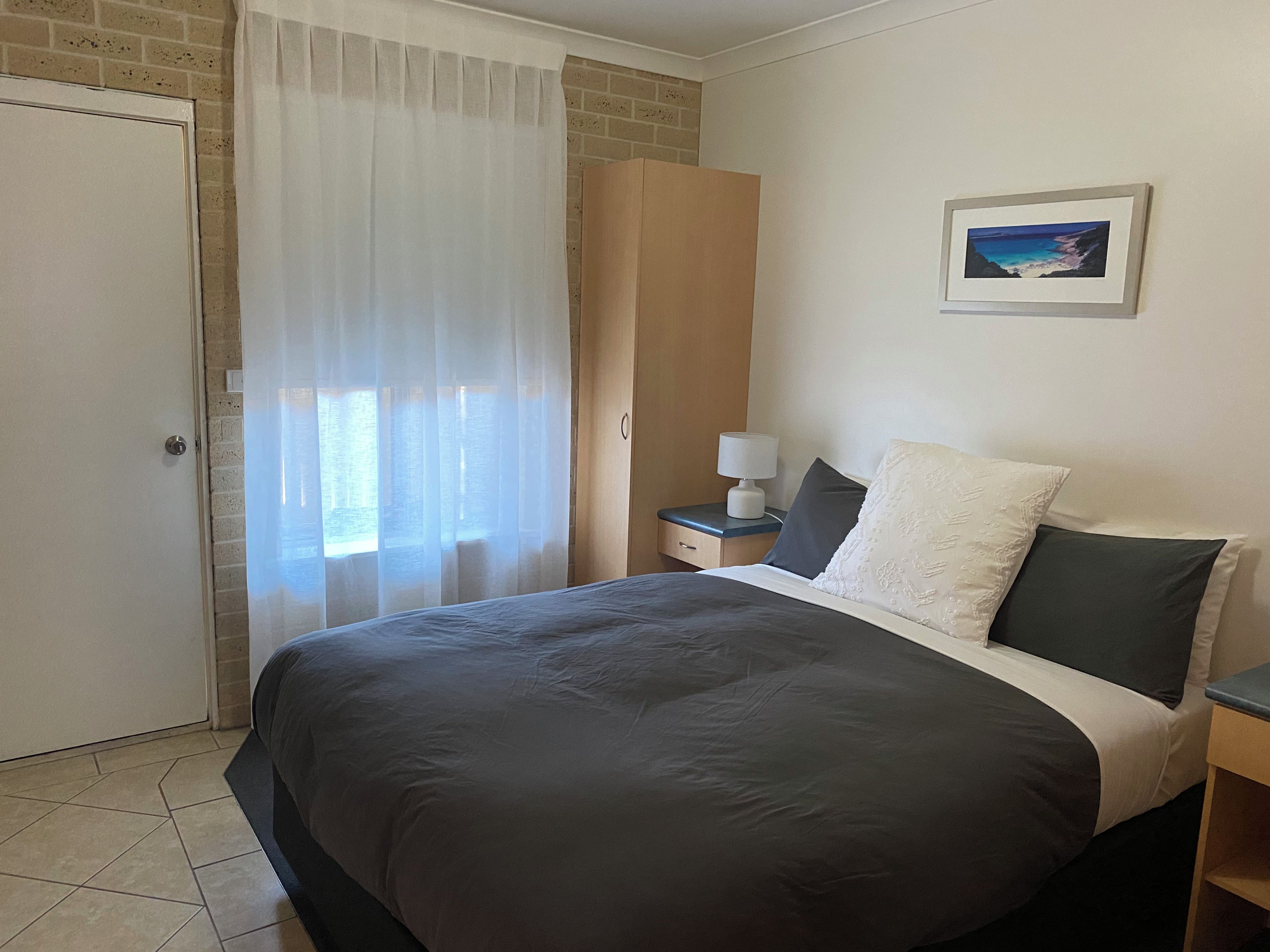 A hotel room with a bed, a nightstand, a lamp, a framed picture on the wall, and a window with sheer curtains.