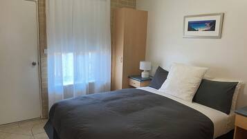 A hotel room with a bed, a nightstand, a lamp, a framed picture on the wall, and a window with sheer curtains.