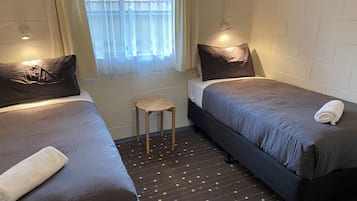 Ein Zimmer mit zwei Betten, einem kleinen Hocker und einem Fenster mit Vorhängen.