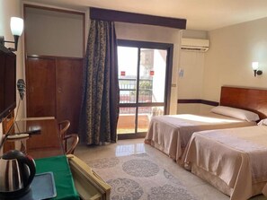 A hotel room with two beds, a desk with a chair, a green table, and a view of the outside. - Stjosephhotel (Luxor)