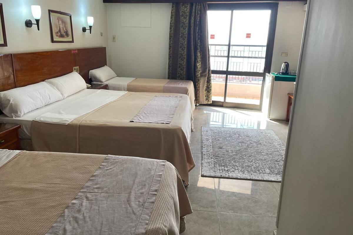 A hotel room with three beds, a small desk, and a view of the outside.