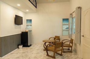 Una habitación con televisión montada en la pared, una pequeña mesa con sillas, un cubo de basura y una ventana con cortinas.