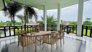 A covered outdoor seating area with a table and chairs, surrounded by a black railing, overlooking a green landscape with palm trees. - Saithong Resort (Pranburi)