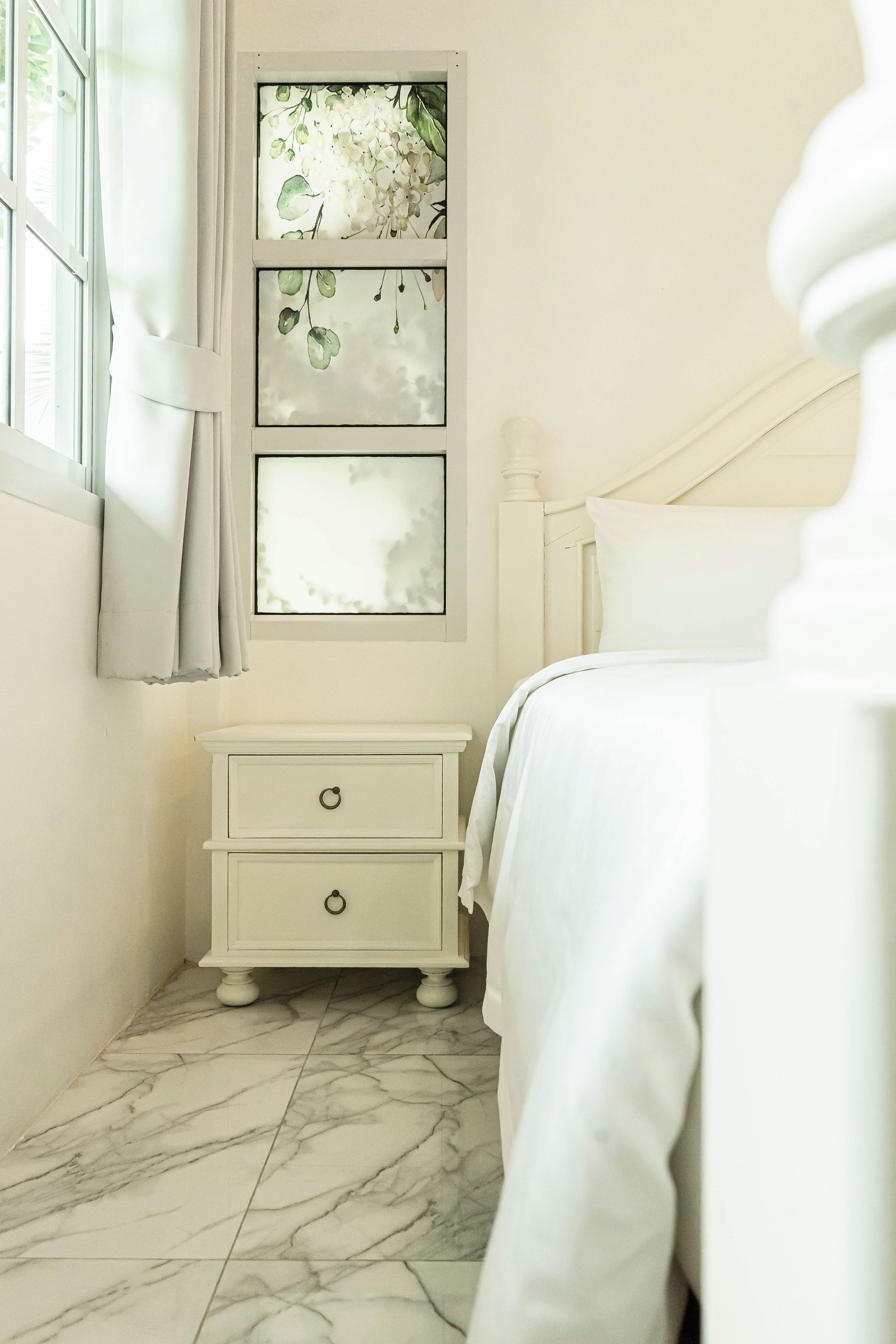 A bedroom with a bed, a nightstand, a window with curtains, and a wall-mounted artwork.